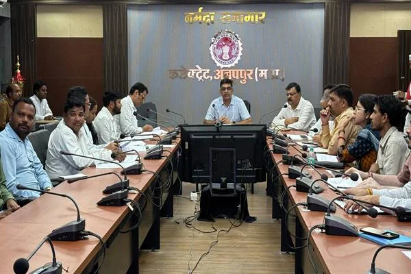 कलेक्ट्रेट अनूपपुर के नर्मदा सभागार में यू-आकार की मेज पर बैठे अधिकारी, सामने माइक और फाइलें; मुख्य कुर्सी पर बैठे अधिकारी बैठक ले रहे हैं, पीछे दीवार पर 'नर्मदा सभागार, कलेक्ट्रेट अनूपपुर (म.प्र.)' लिखा है।