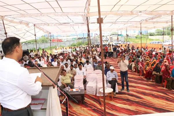 मंच से संबोधित करते अधिकारी के सामने पंडाल में बैठे ग्रामीण, महिलाएं और अन्य उपस्थित लोग