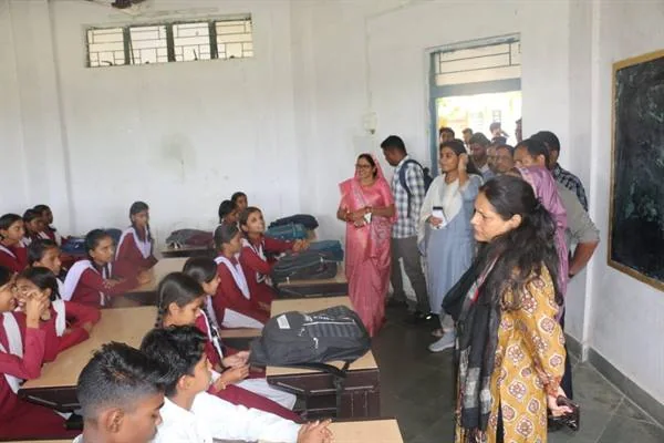 कक्षा में डेस्क पर बैठे विद्यार्थियों से बातचीत करते प्रशासनिक अधिकारी और अन्य लोग