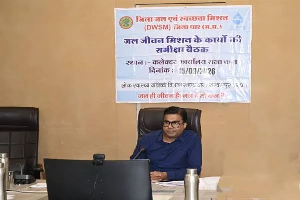 बैठक कक्ष में मेज के पीछे बैठे अधिकारी और सामने रखा कंप्यूटर मॉनिटर, पीछे जिला जल एवं स्वच्छता मिशन की समीक्षा बैठक का बैनर
