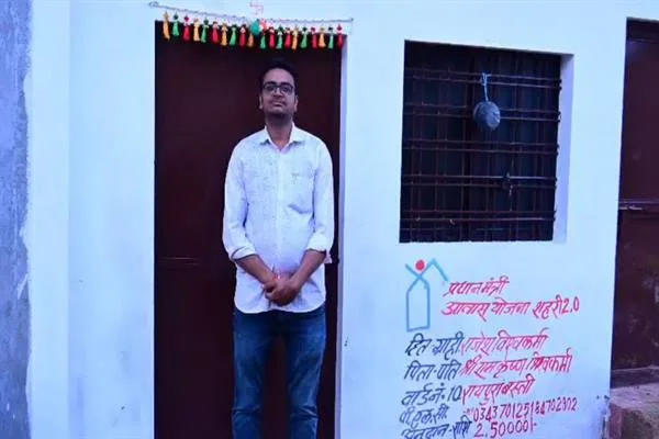 पक्के मकान के दरवाजे पर खड़ा चश्मा लगाए एक व्यक्ति, पास की दीवार पर आवास योजना का विवरण लिखा है
