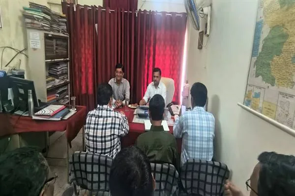 कार्यालय के एक कमरे में मेज के सामने कुर्सियों पर बैठे लोगों के साथ बैठक करते दो अधिकारी