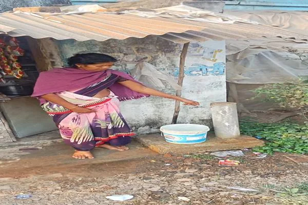 एक महिला गांव में कच्चे आंगन के पास बैठकर दीवार से लगे नल से पानी भर रही है, पास में प्लास्टिक की बाल्टी रखी है