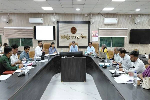 कलेक्ट्रेट के वातानुकूलित सभाकक्ष में यू-आकार की बड़ी मेज के चारों ओर बैठे करीब बीस अधिकारी, सामने कुर्सी पर बैठे अध्यक्षता कर रहे अधिकारी, मेज पर फाइलें, कागज और पानी की बोतलें; पीछे दीवार पर कार्यालय का नाम-पट्ट और बाईं ओर स्क्रीन पर प्रस्तुति।